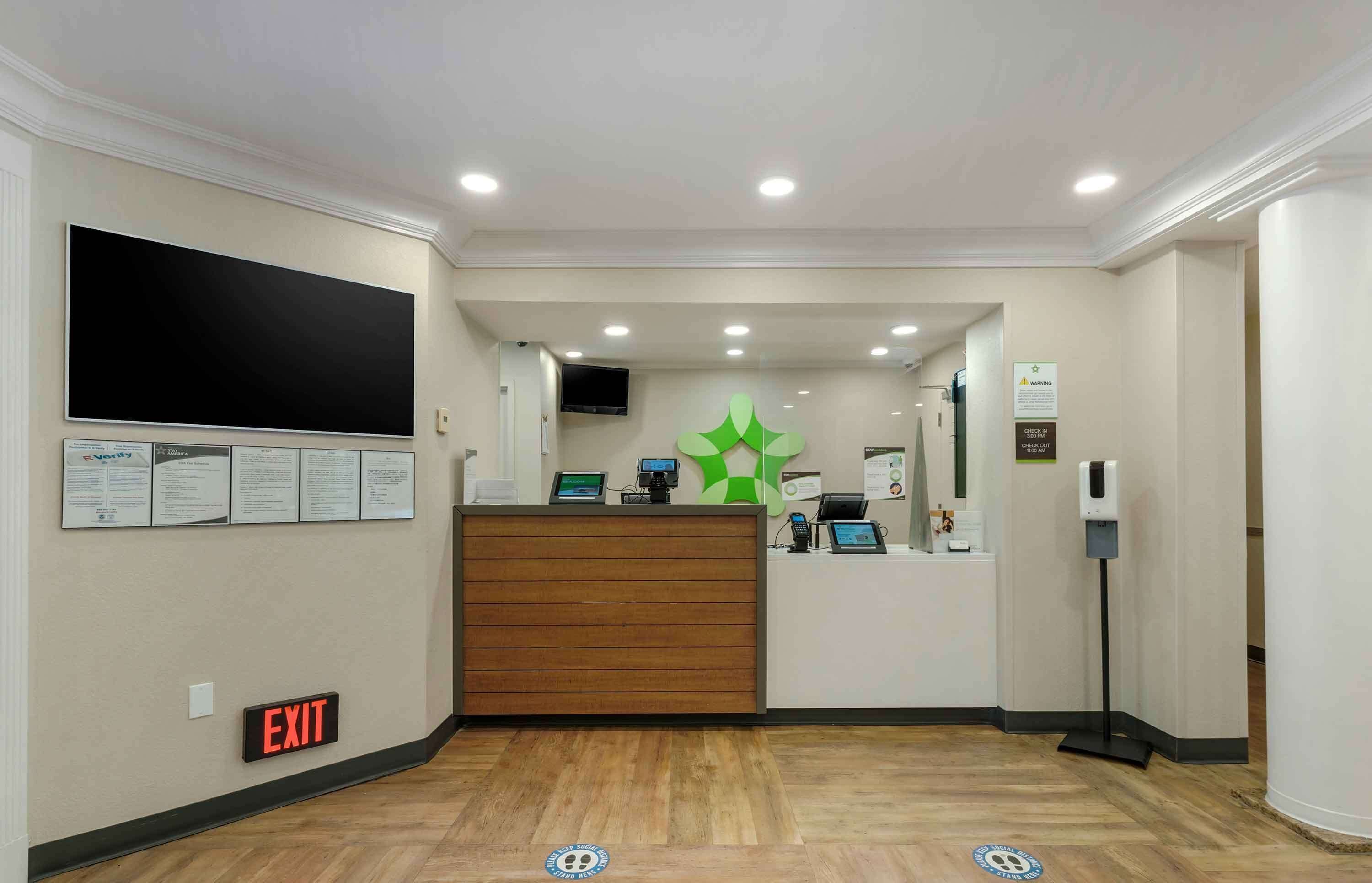 A reception area with a wooden reception desk, a large flat-screen TV, a green logo on the wall, and an exit sign.