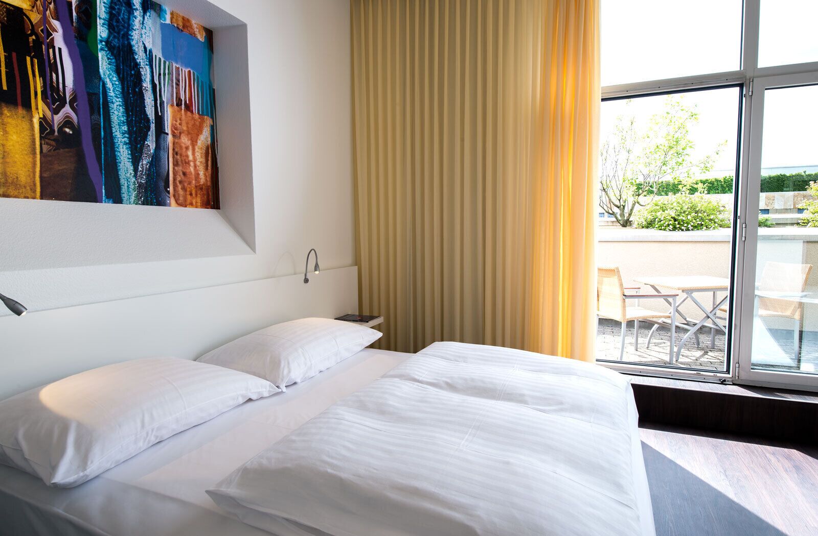 Habitación de hotel con una cama grande, un cuadro en la pared, una ventana con cortinas y vistas al exterior.