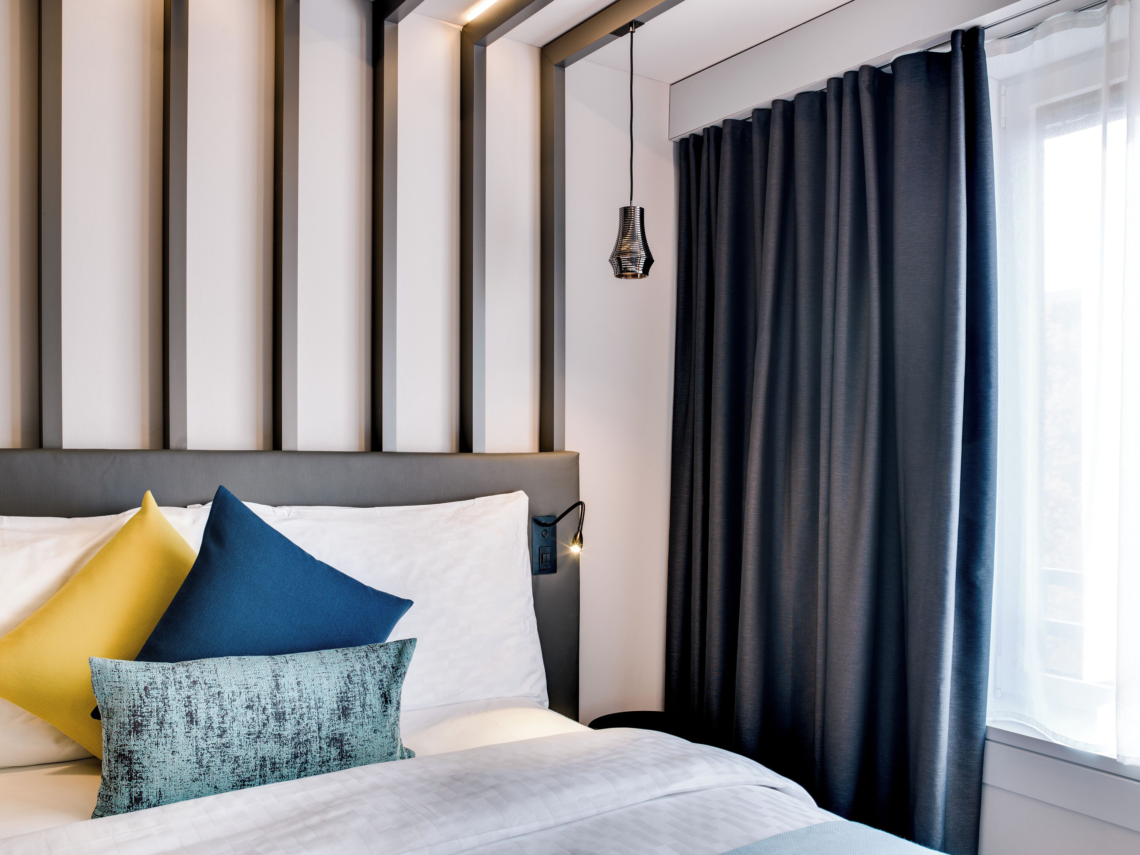 A modern hotel room with a large bed, a headboard with vertical lines, and a window with dark curtains.
