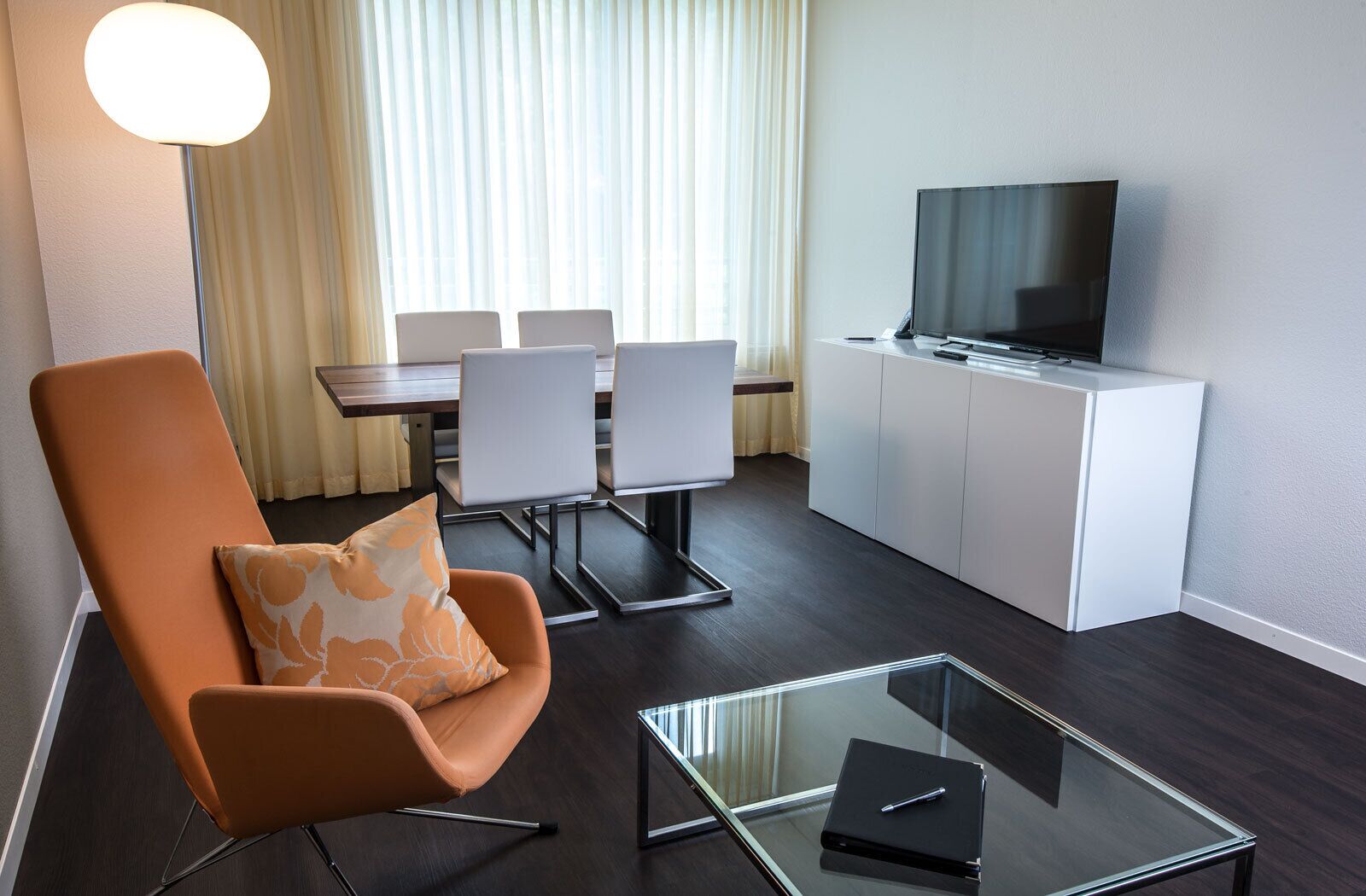 A modern hotel room with a glass coffee table, an orange armchair, a dining area with white chairs, and a flat-screen TV on a white cabinet.