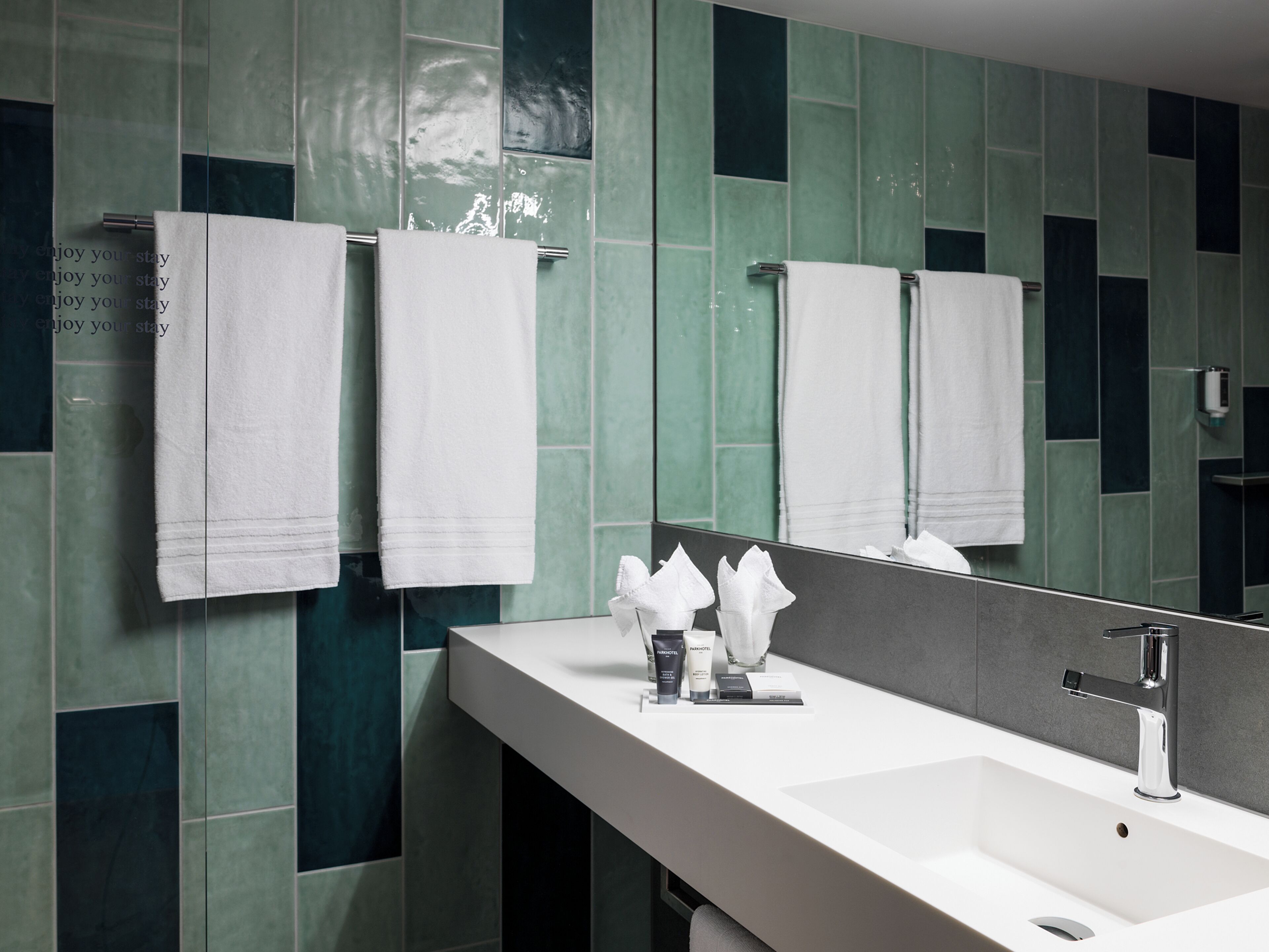 A modern bathroom with a white sink, a mirror, and green tiled walls.
