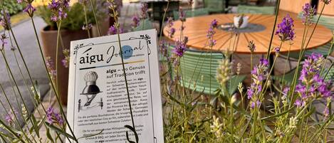 A menu titled 'aigu Journal' with text in German, surrounded by purple flowers and green plants, set against a backdrop of outdoor seating with tables and chairs.