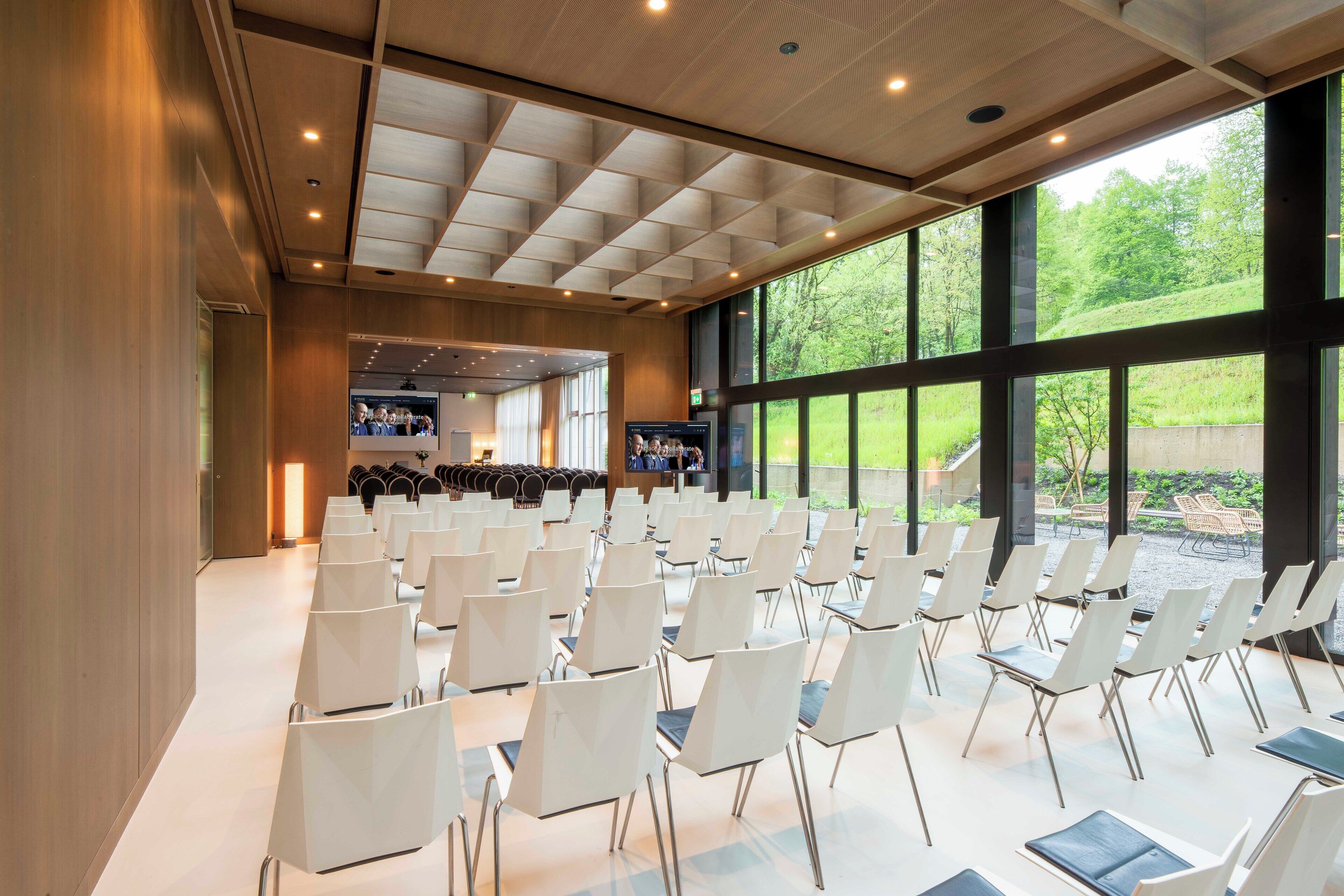 Ein Konferenzraum mit Reihen weißer Stühle, großen Fenstern und einer Holzbalkendecke.