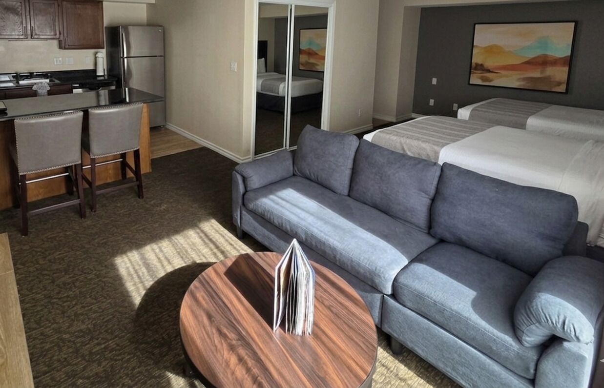 Une chambre d’hôtel avec une kitchenette, un canapé gris, une table basse ronde en bois et un lit avec du linge de maison blanc.