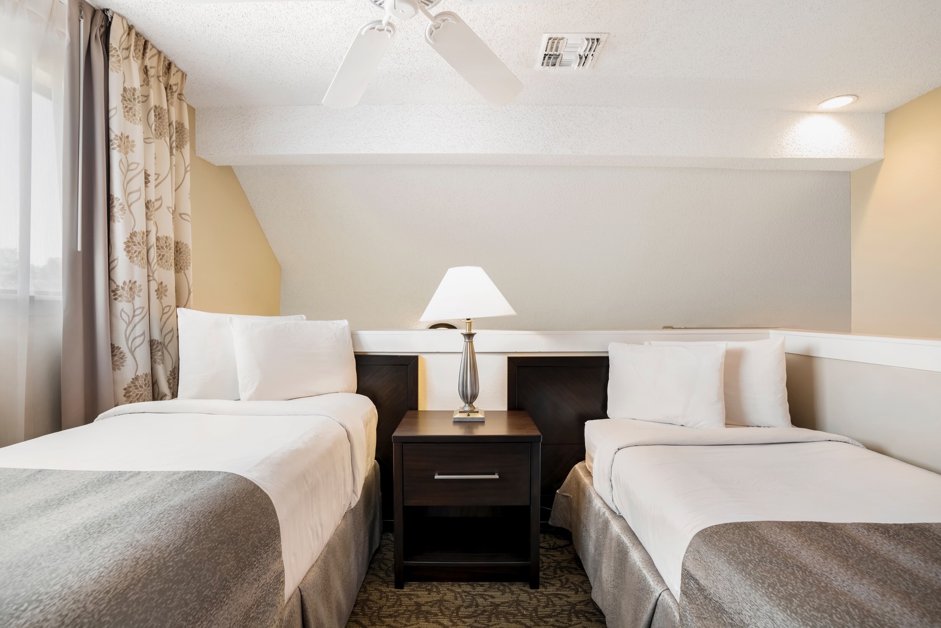 Ein Hotelzimmer mit zwei Betten, einem Nachttisch mit Lampe, einem Deckenventilator und einem Fenster mit Vorhängen.