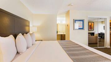 A hotel room with a large bed, a dark headboard, a bedside lamp, a mirror, and a door leading to a bathroom and a sitting area.