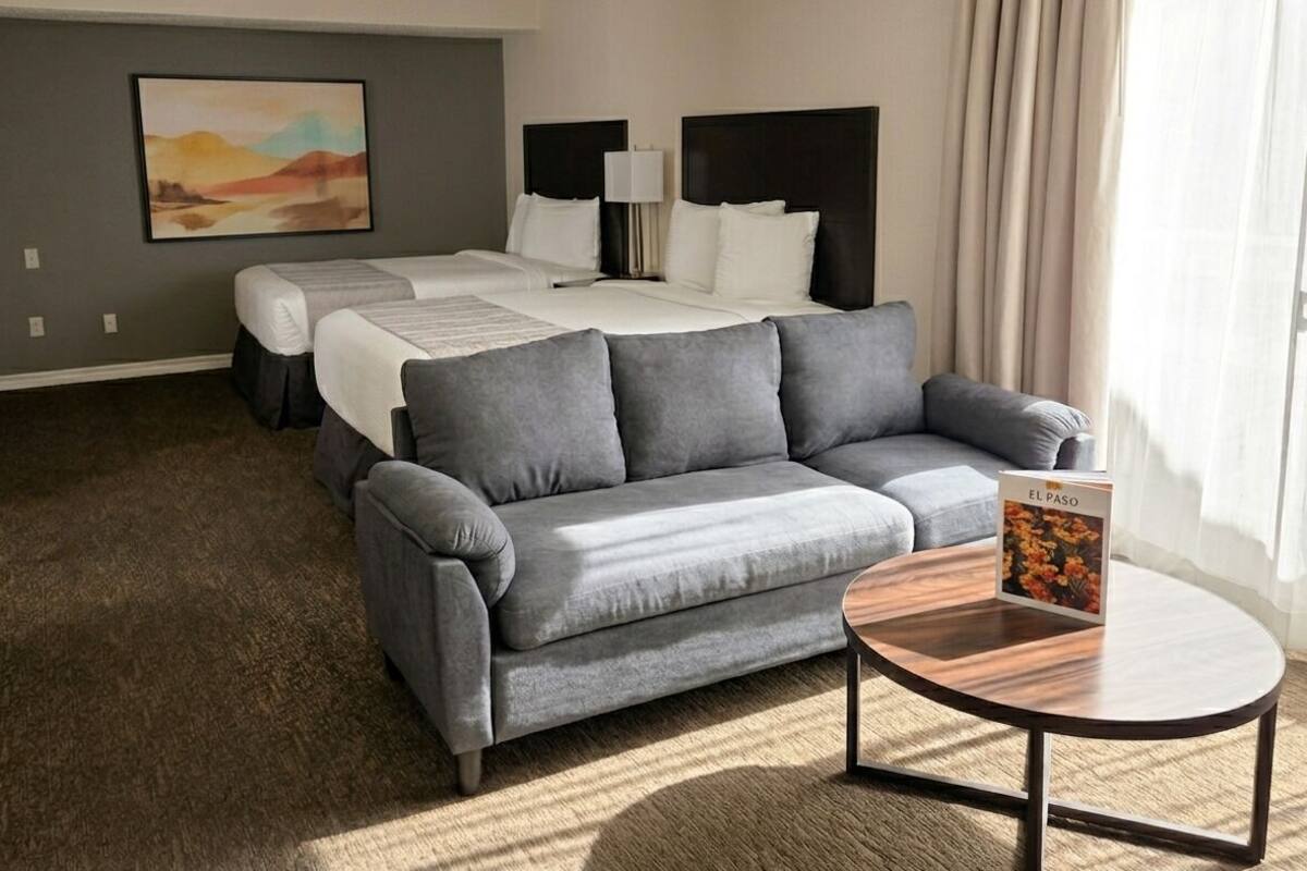Quarto de hotel com cama, sofá cinzento, mesa redonda pequena e um quadro na parede.