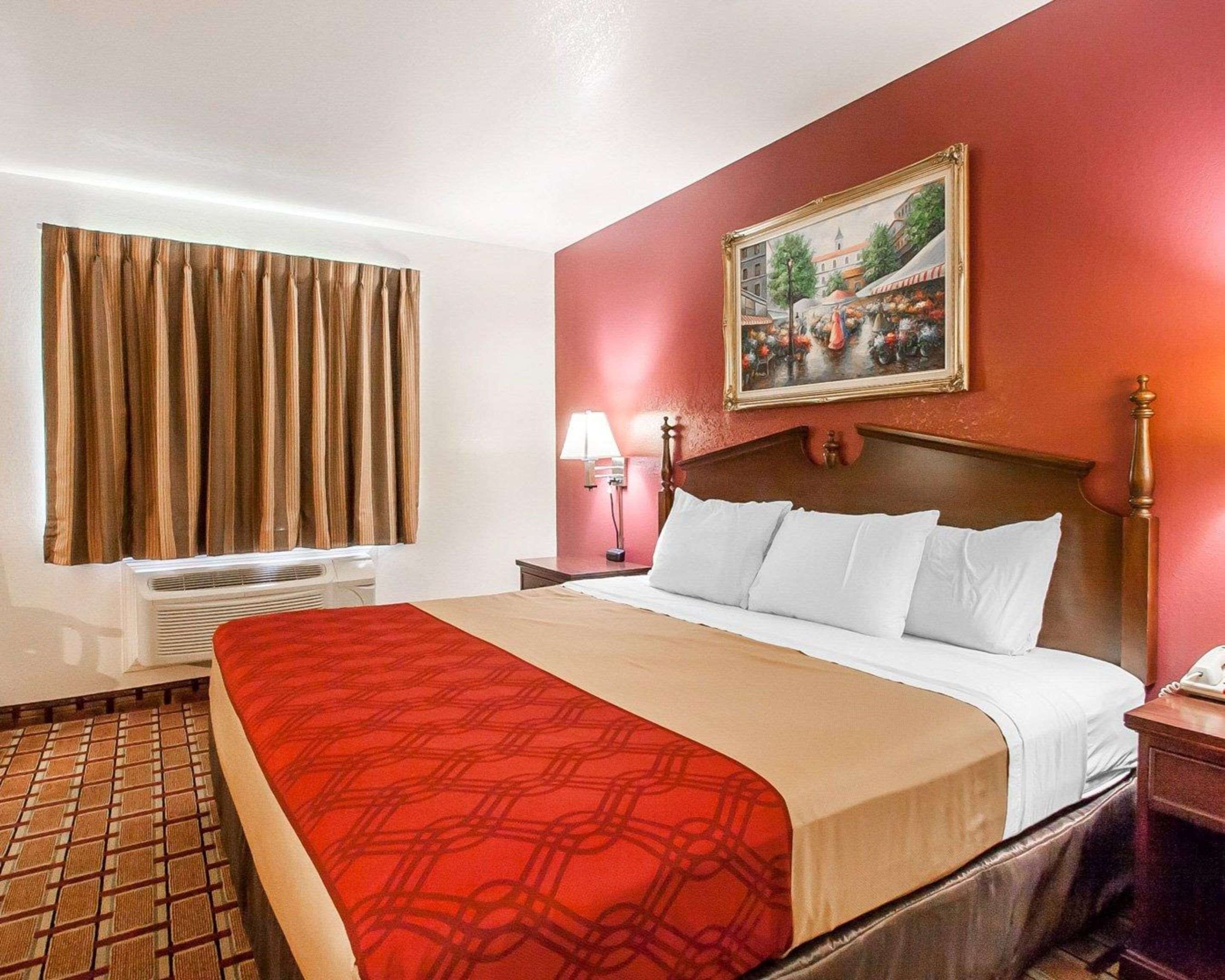 Habitació d'hotel amb un llit gran, un quadre a la paret, una taula de treball amb una làmpada i una finestra amb cortines.