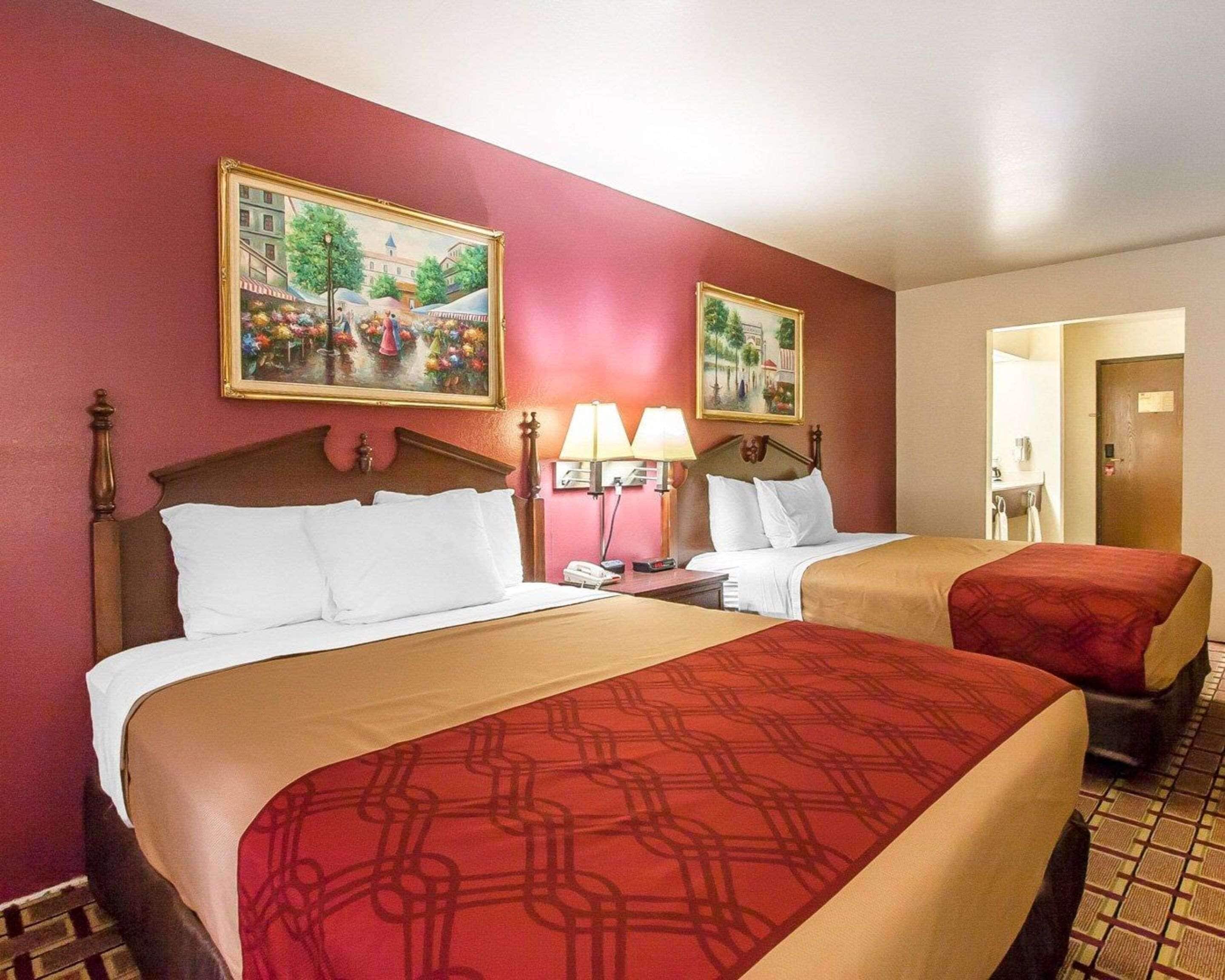 A hotel room with two beds, each with a red and beige bedspread, a wooden headboard, and a nightstand with a lamp. There are two framed paintings on the wall, and a door leading to another room.