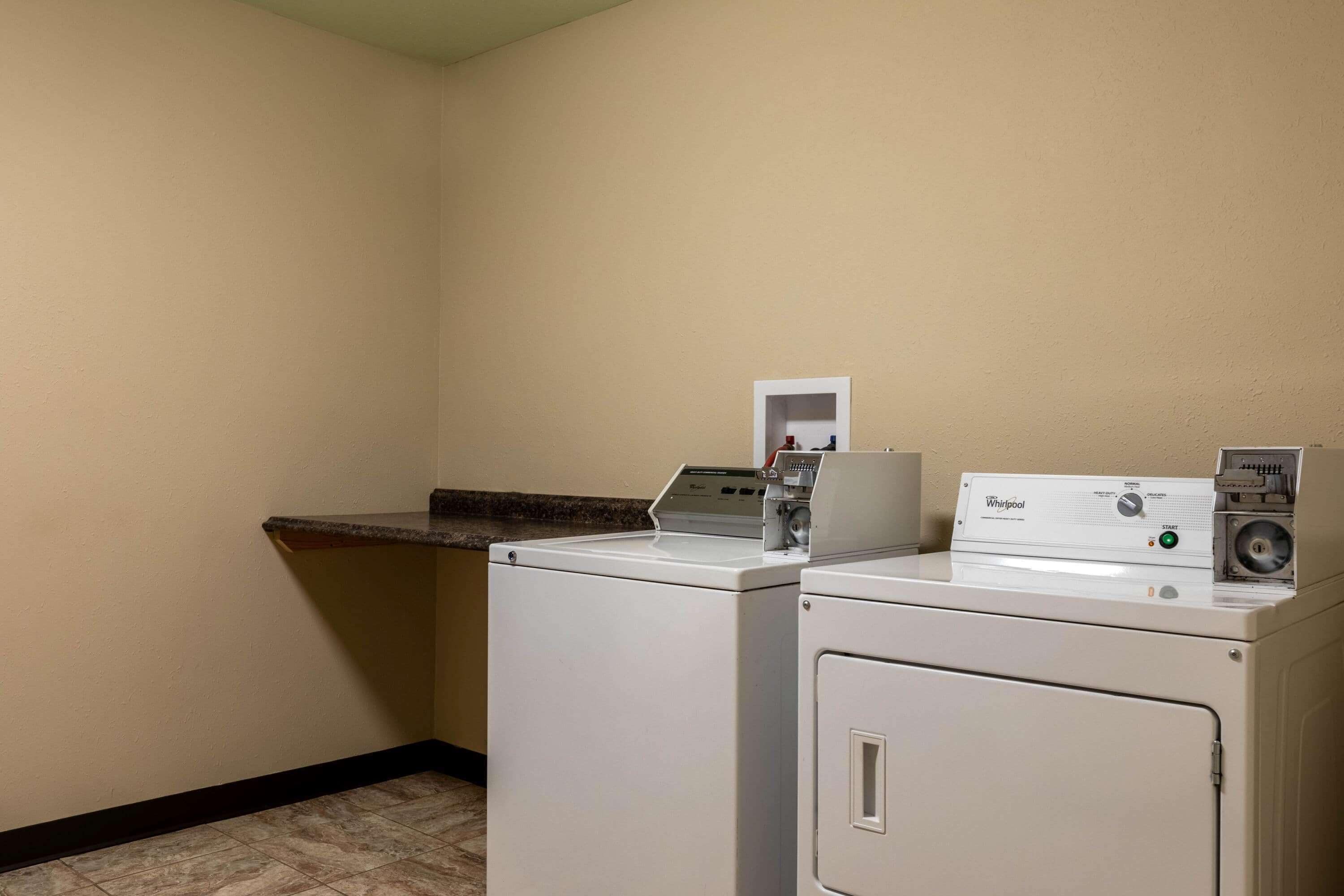 A laundry room with two washers and a dryer, a small shelf, and a wall-mounted electrical outlet.