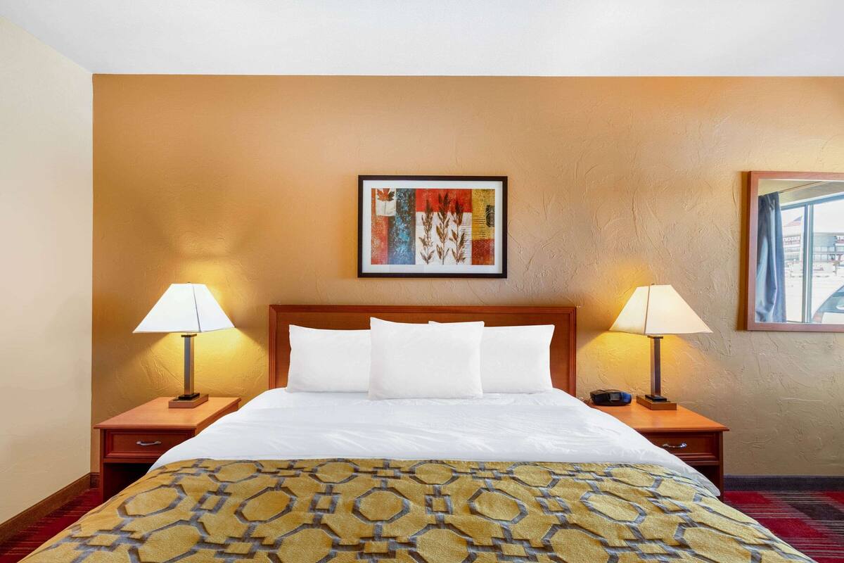 Een hotelkamer met een groot bed, twee nachtkastjes met lampen, een kunstwerk in een lijst boven het bed, en een raam met uitzicht.
