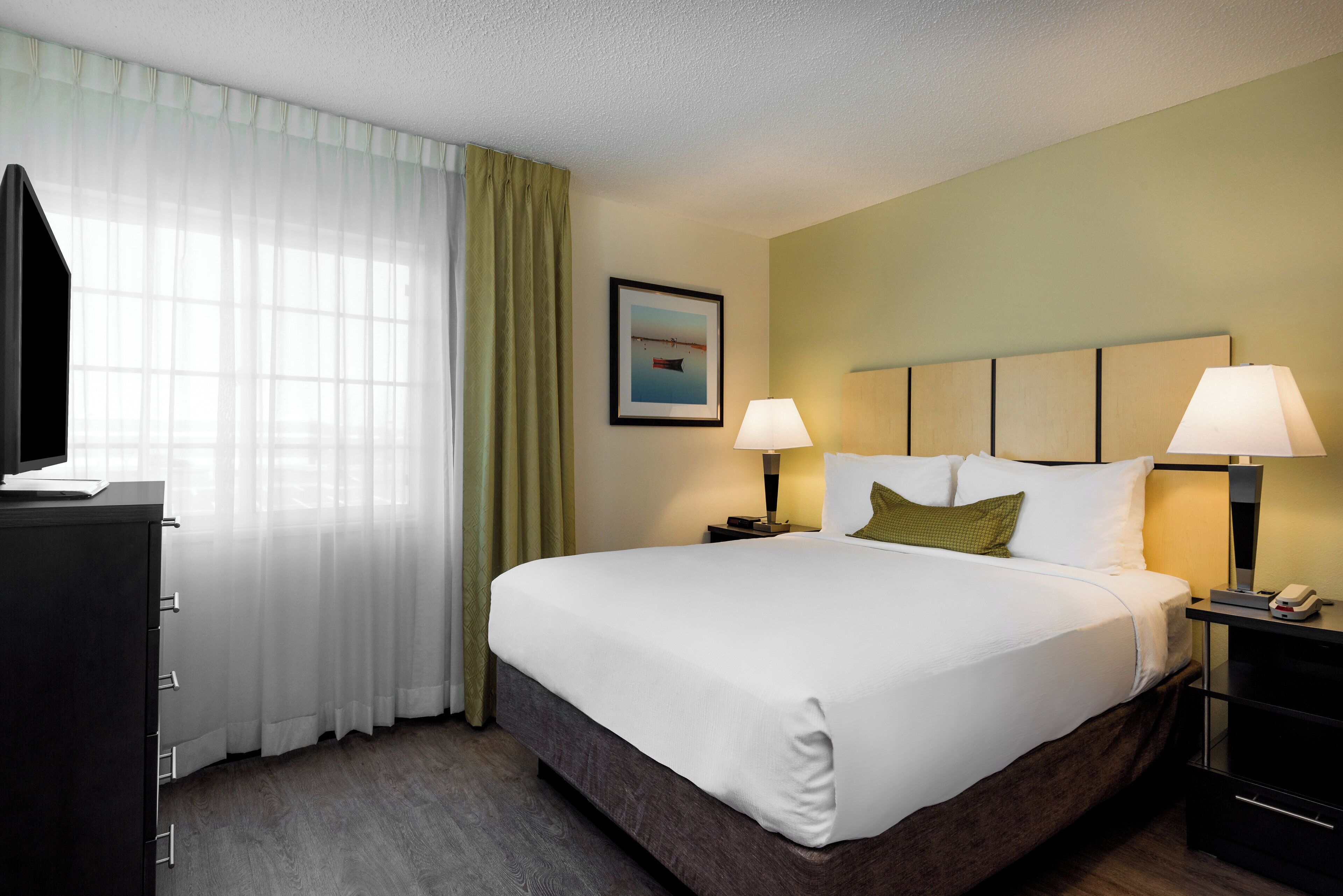 A hotel room with a large bed, two bedside tables with lamps, a flat-screen TV, a window with sheer curtains, and a framed picture on the wall.