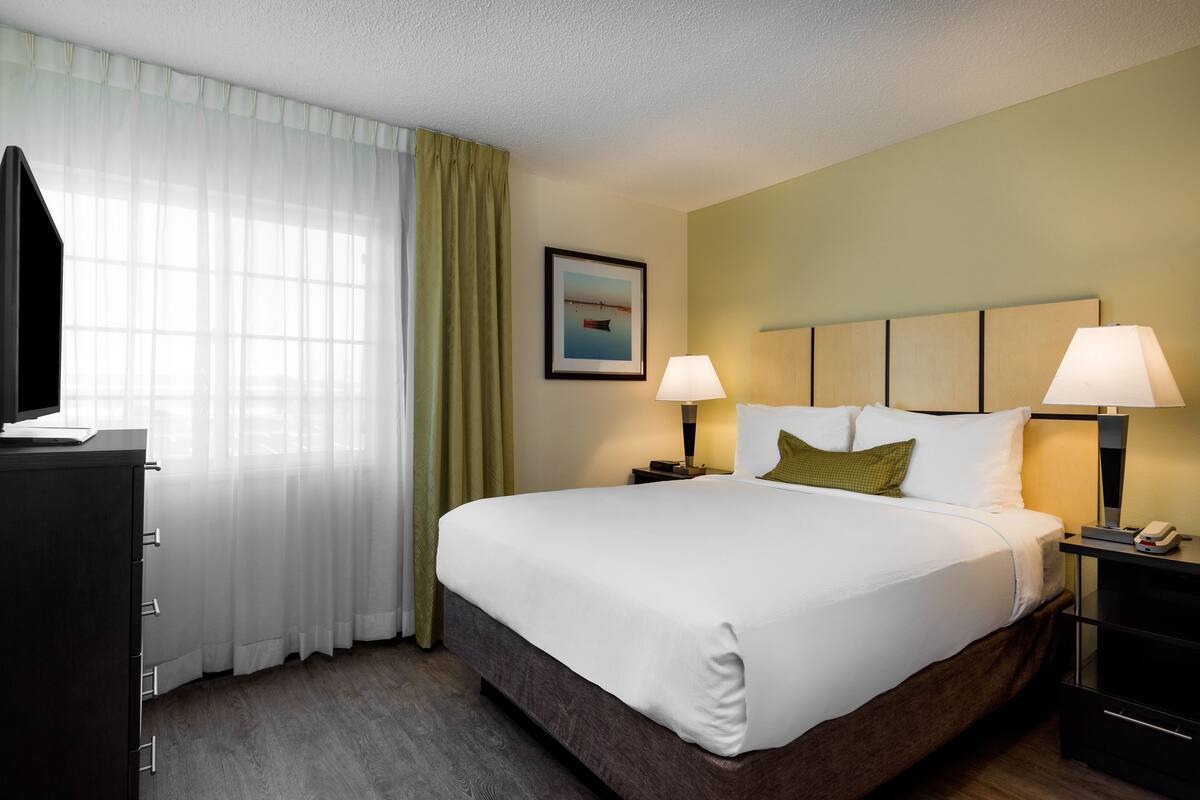 Habitación de hotel con una cama grande, dos lámparas de noche, una mesita de noche, un escritorio con teléfono, un televisor y una ventana con cortinas translúcidas.