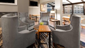 A modern hotel room with grey upholstered chairs, a wooden coffee table, and a large window offering a view of buildings.