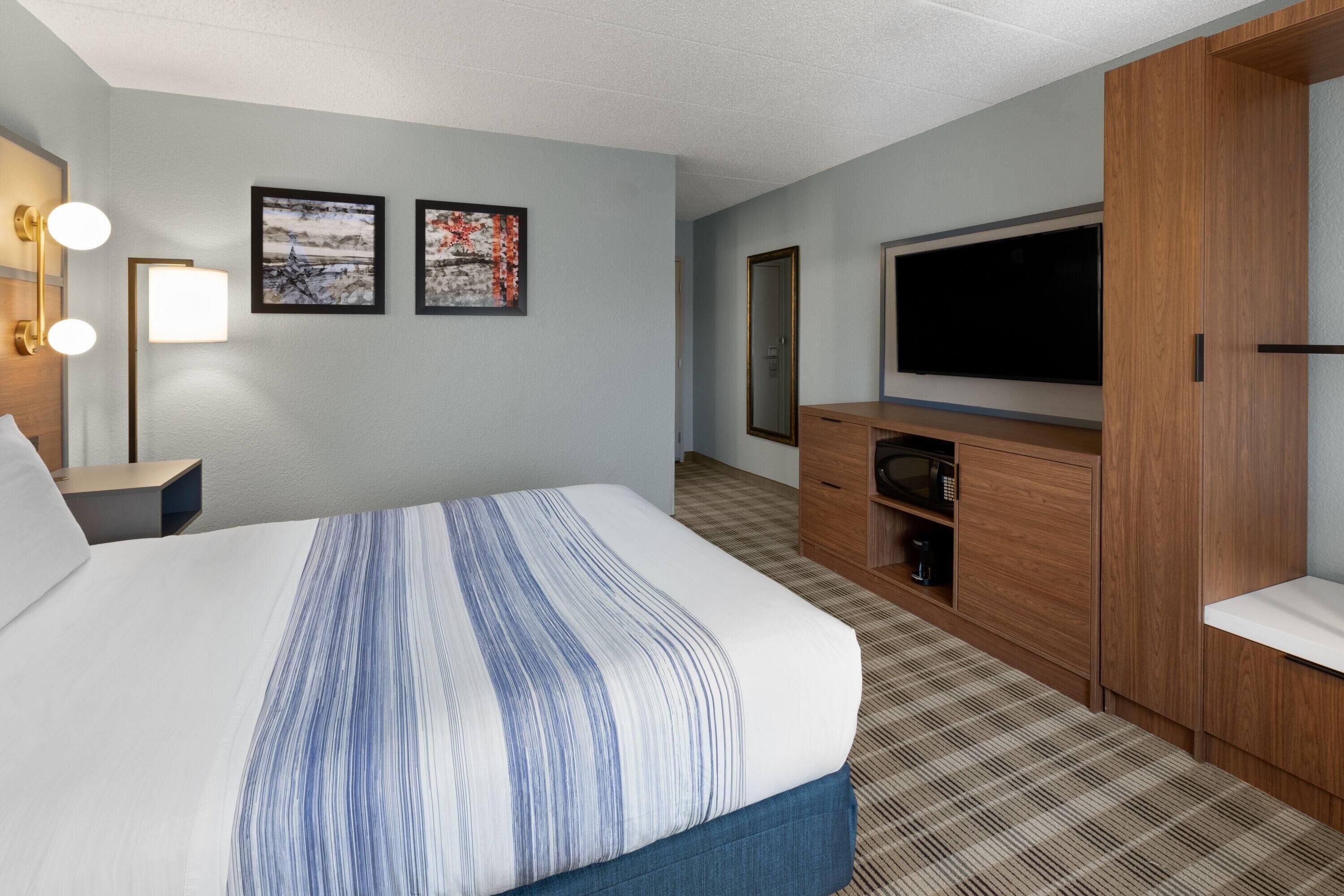 A hotel room with a large bed, a flat-screen TV, a wooden wardrobe, a desk with a lamp, and two framed pictures on the wall.
