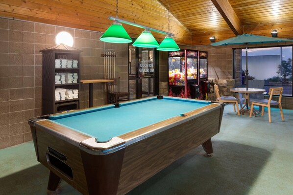 A pool table with three hanging lamps, a pool cue rack, and a seating area with chairs and a table.