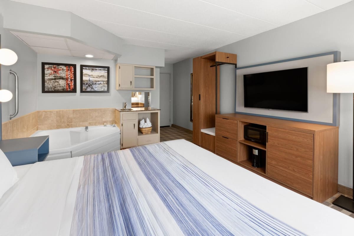 A modern hotel room with a large bed, a built-in TV, a wooden cabinet, a bathtub, and a small kitchenette.