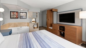 A modern hotel room with a large bed, a built-in TV, a wooden cabinet, a bathtub, and a small kitchenette.