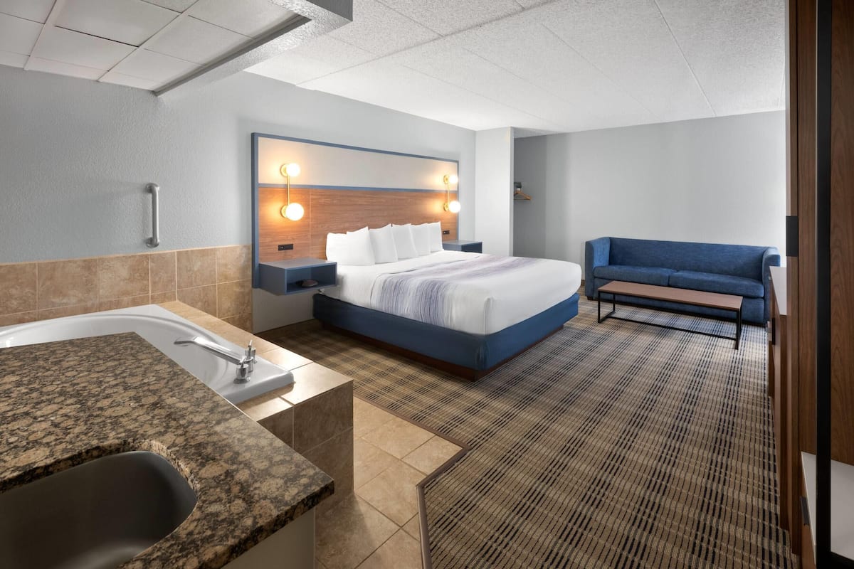 A hotel room with a bed, a bench, a bathroom with a sink and bathtub, and a wall-mounted light fixture.