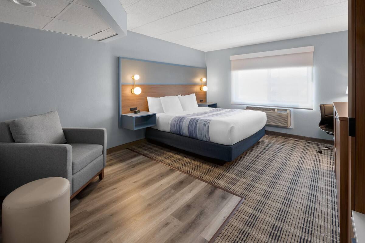 A modern hotel room with a large bed, a grey armchair, a small round ottoman, and a desk with a chair.