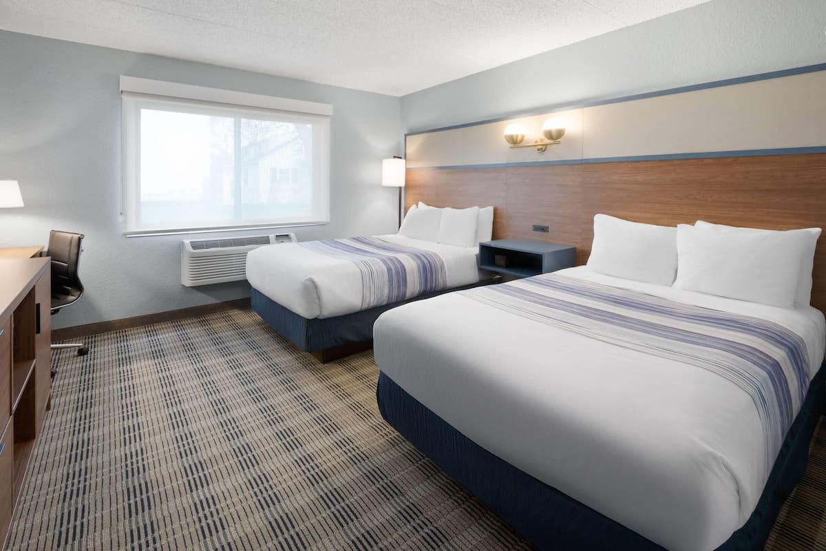 A hotel room with two beds, a desk, a chair, a lamp, and a window.