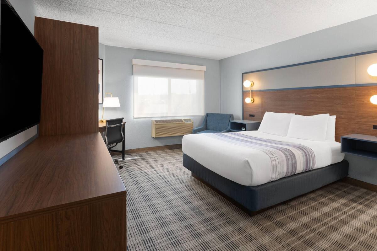A hotel room with a large bed, a desk, a television, and a window with blinds.