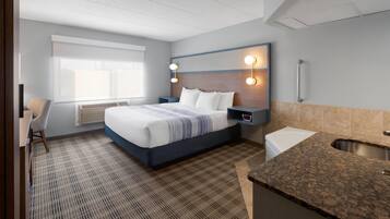 A hotel room with a bed, bedside table, lamp, and a small kitchenette with a sink.