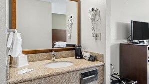 A hotel bathroom with a granite countertop sink, a large mirror, a hairdryer, a television, and a cabinet.