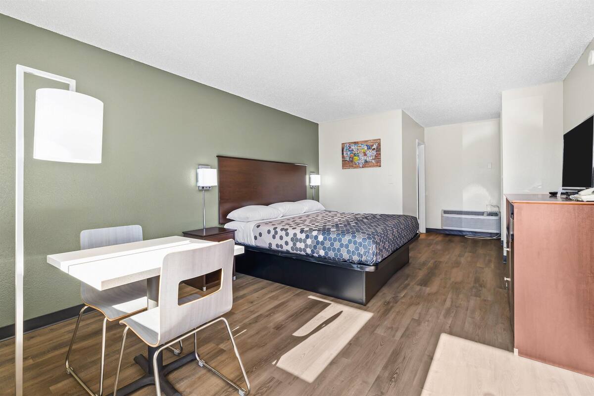 A modern hotel room with a bed, desk, chair, and a small kitchenette.