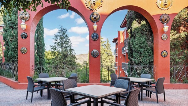 Un espace extérieur aménagé en terrasse, avec des tables et des chaises, entouré d’arcs rouges ornés de motifs solaires décoratifs.