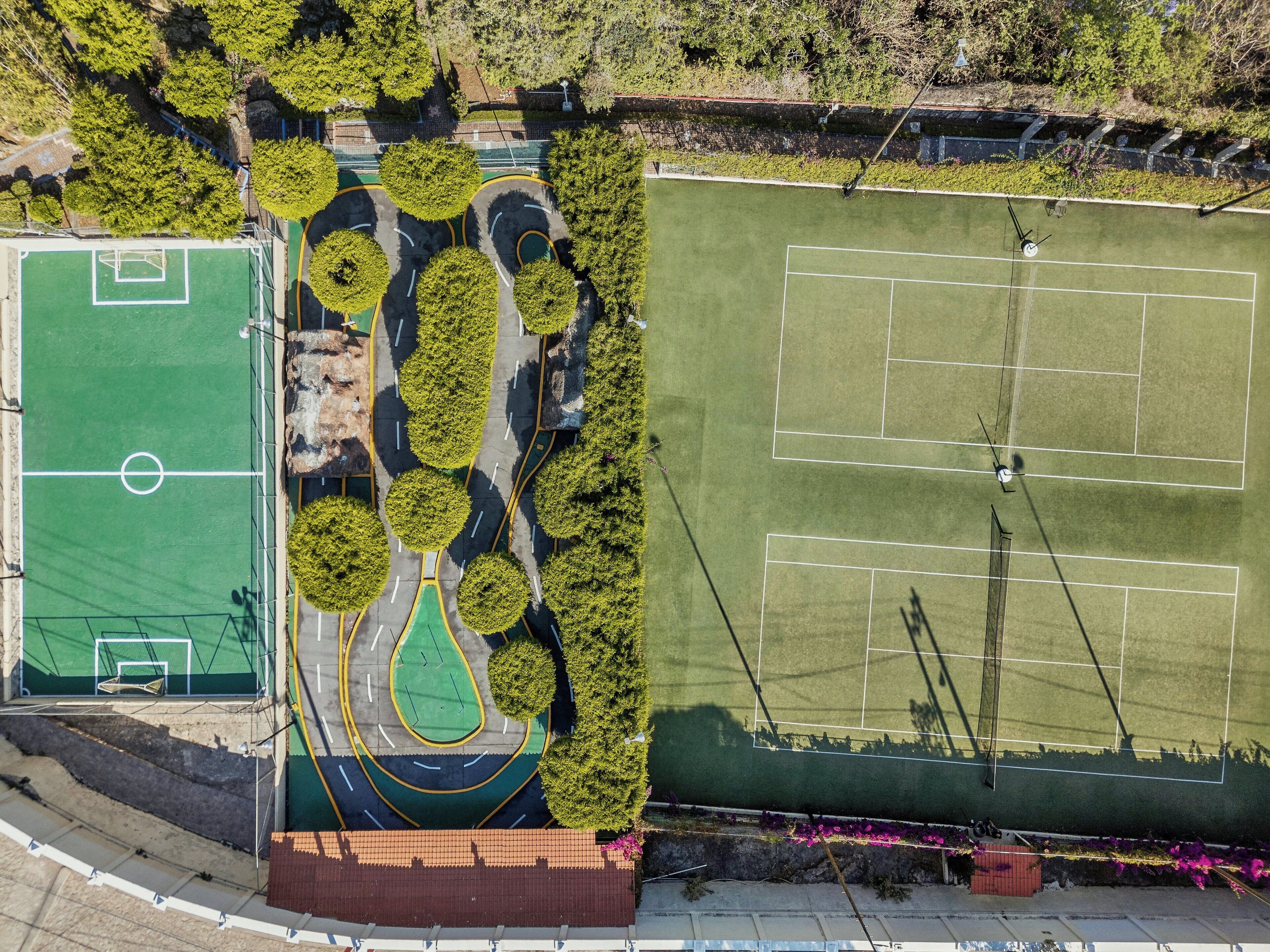 Vista aérea de un complejo deportivo con varias canchas, incluyendo un campo de fútbol y canchas de tenis, rodeado de árboles y senderos.