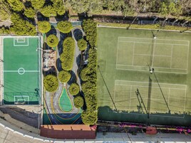 Vista aérea de un complejo deportivo con varias canchas, incluyendo un campo de fútbol y canchas de tenis, rodeado de árboles y senderos.