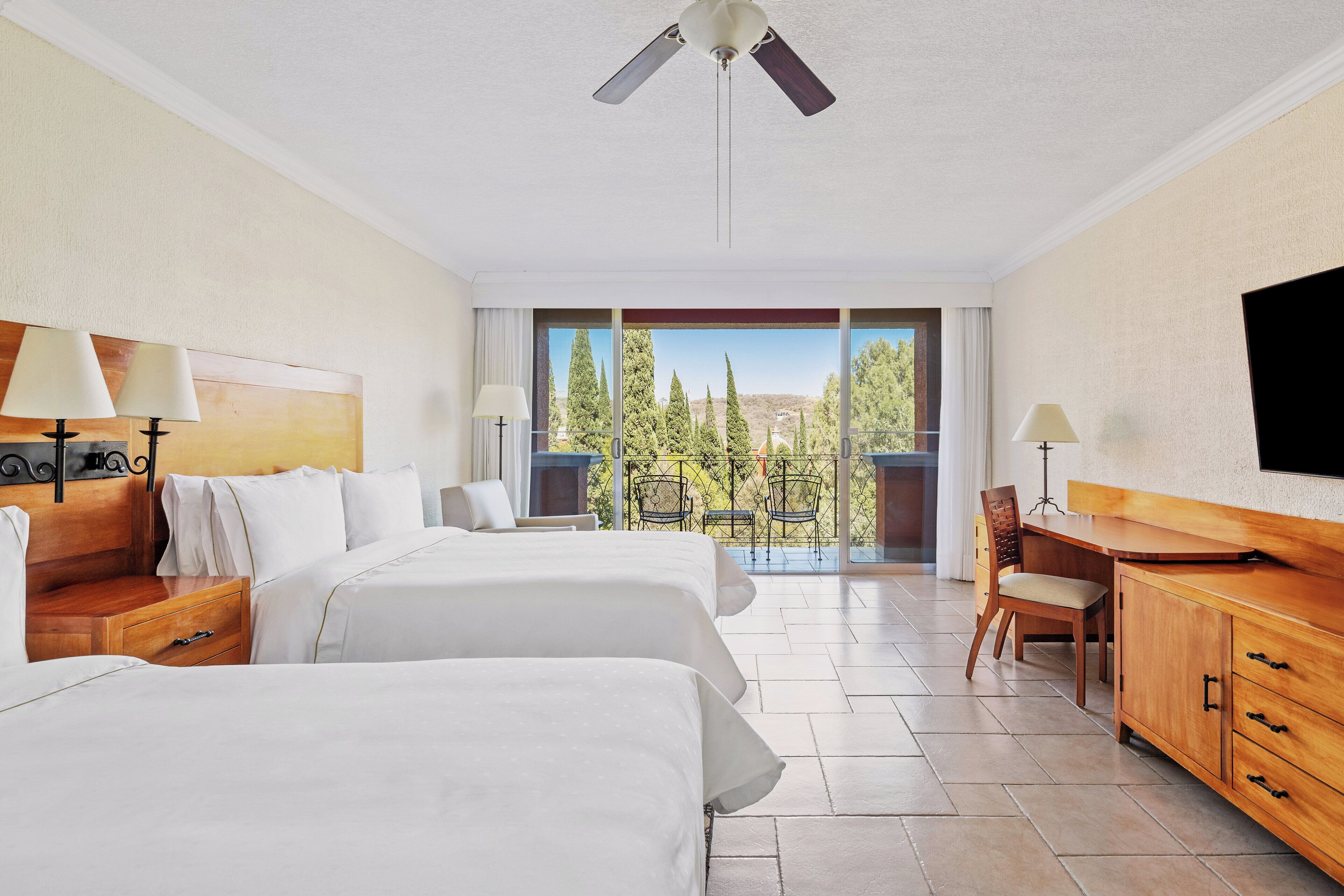 Habitación de hotel con dos camas, un escritorio, una silla, un televisor y un balcón con vistas a árboles.