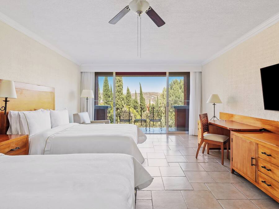 Habitación de hotel con dos camas, un escritorio, una silla, un televisor y un balcón con vistas a árboles.