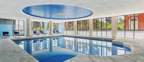 Ein großer, überdachter Swimmingpool mit klarem, blauem Wasser, umgeben von Liegestühlen und großen Fenstern mit Blick ins Freie.