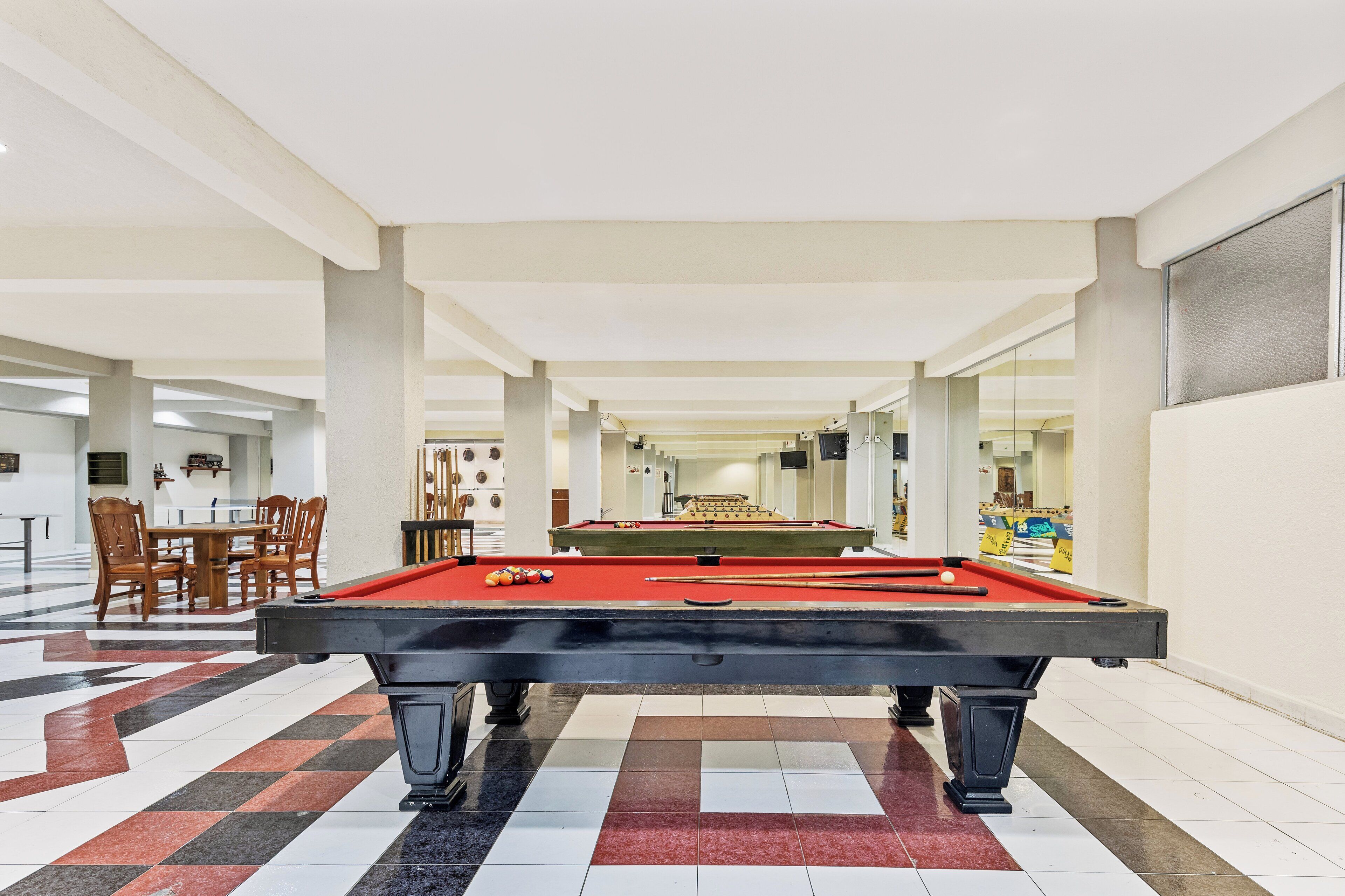 A pool table in a spacious indoor area with a checkered floor, wooden chairs, and a dining table in the background.