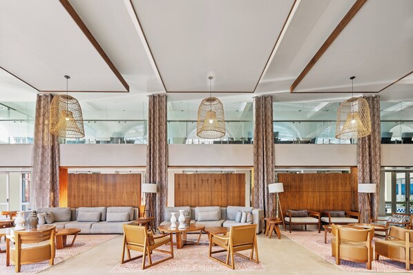 Un hall d’entrée spacieux avec un plafond haut, de grandes fenêtres et un aménagement intérieur moderne comprenant un canapé, des fauteuils et des tables.