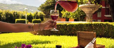 A picnic setup with a wicker basket, a bottle, and a glass of red wine on a blanket in a grassy area with a fountain and trees in the background.