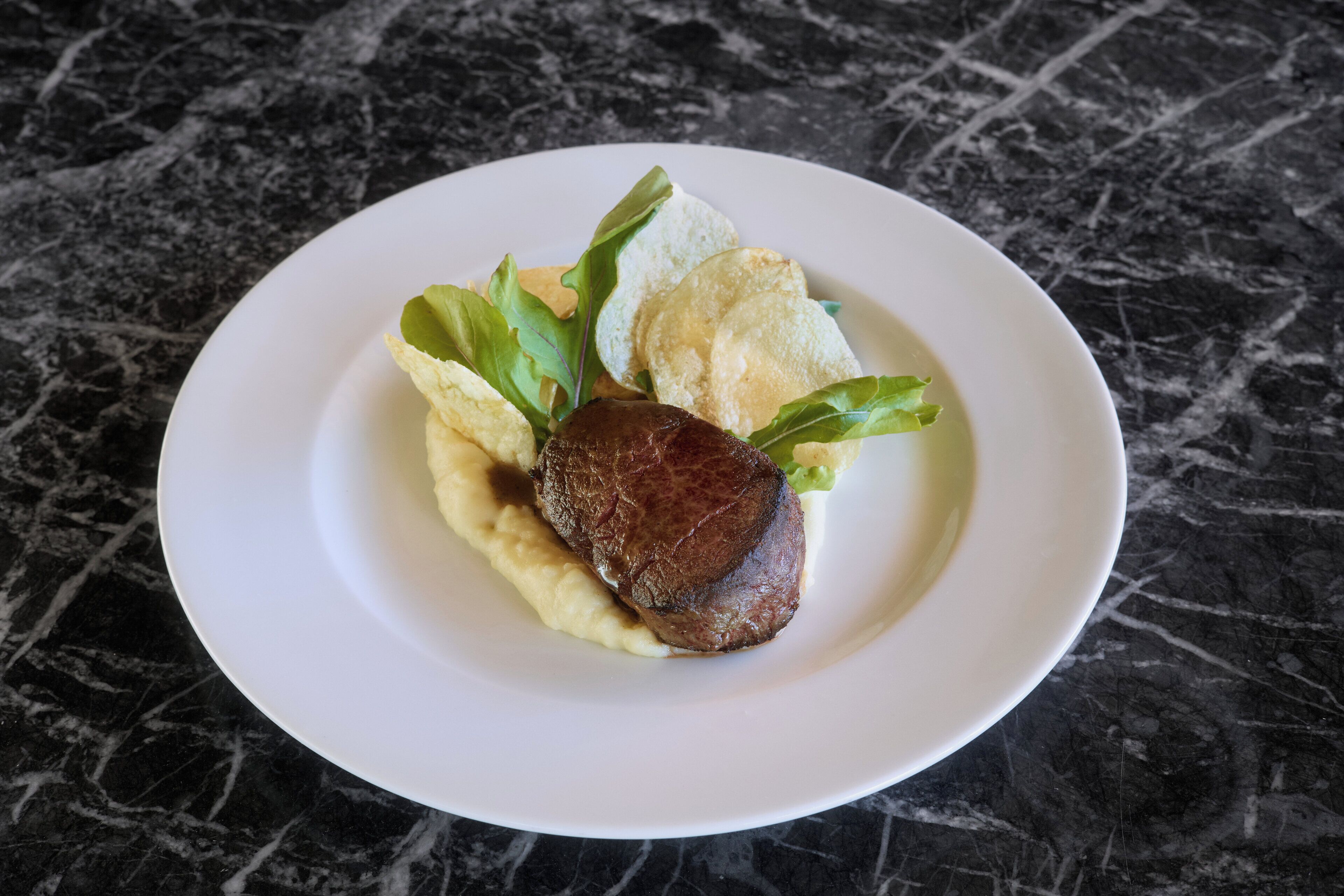 A plate with mashed potatoes, a piece of meat, and some greens on a marble surface.
