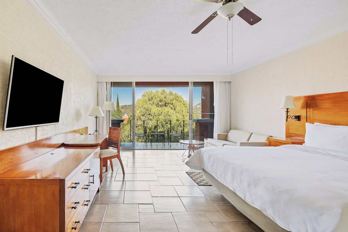 Hotelkamer met een bed, een bureau met een flatscreen-tv, een stoel, een klein tafeltje en een balkon met uitzicht op bomen.