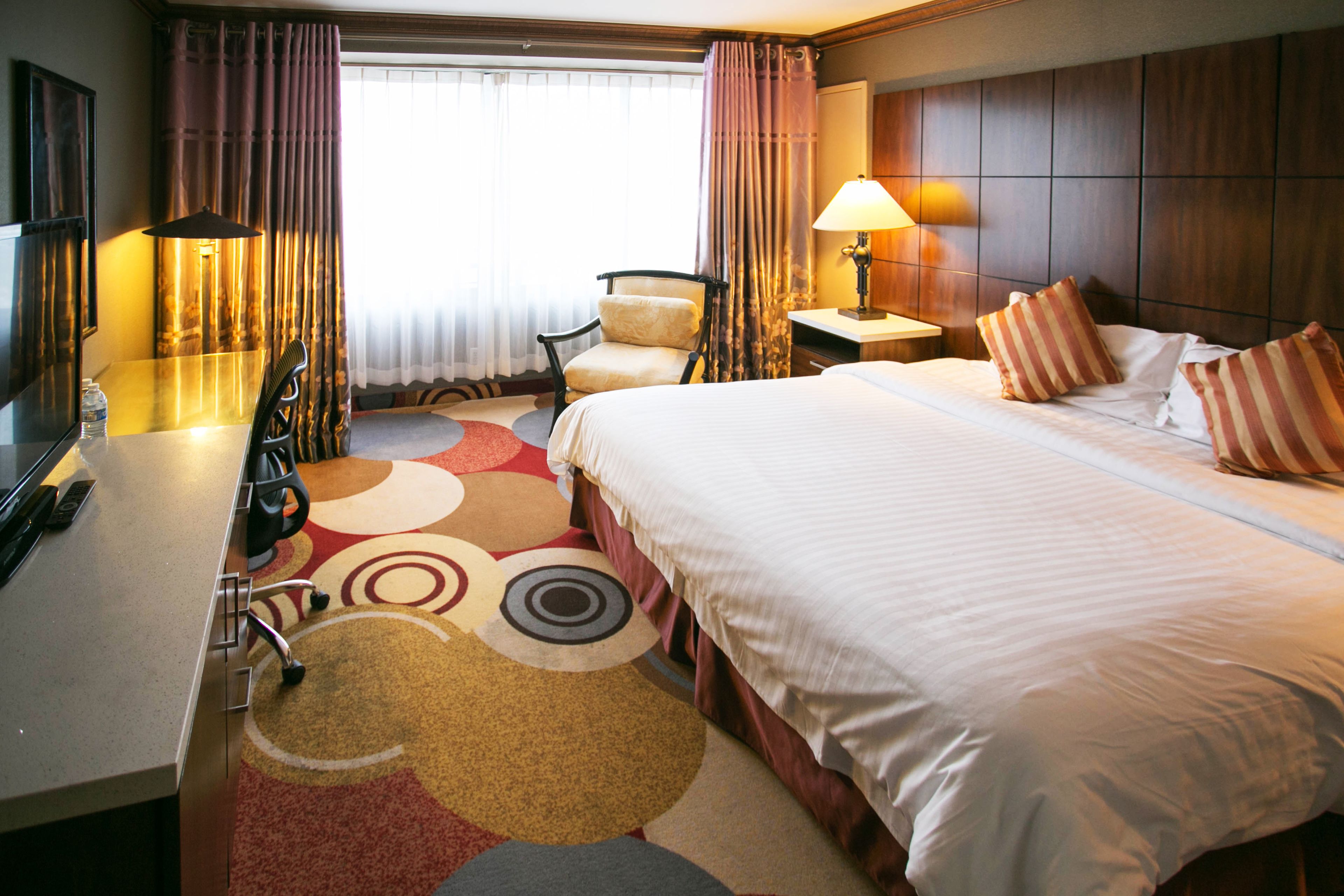 A hotel room with a large bed, a desk with a chair, a lamp, and a view of the outdoors through curtains.