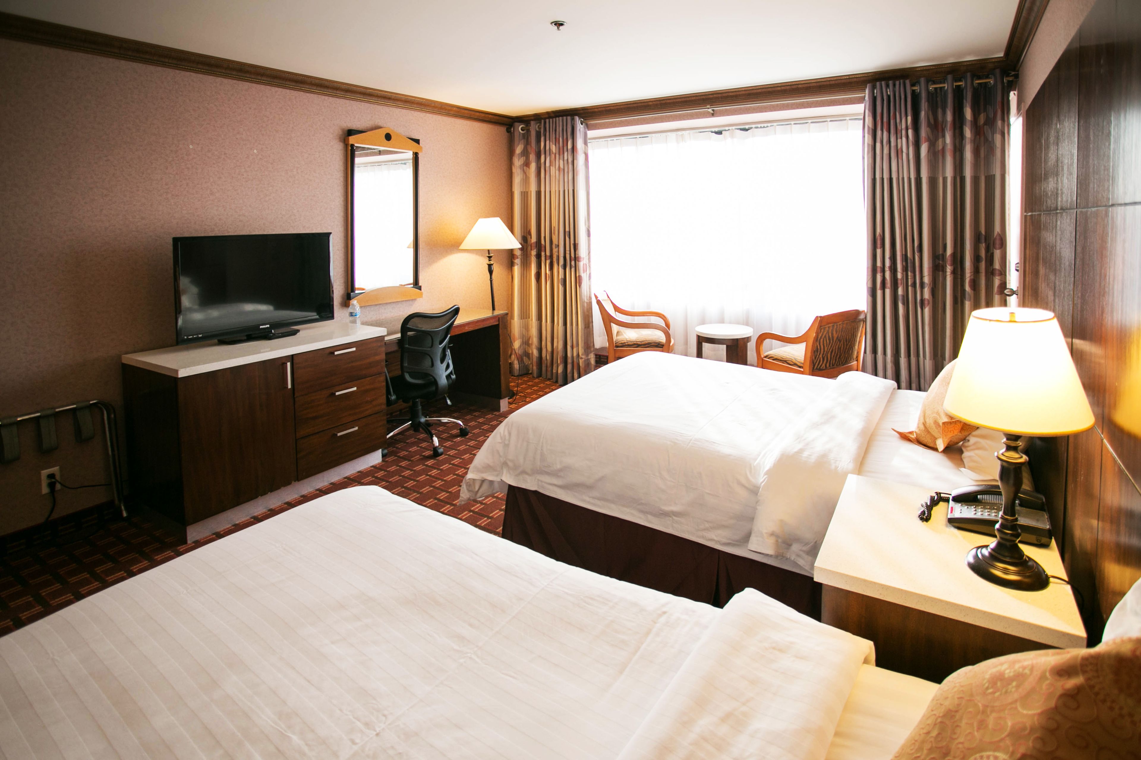 A hotel room with two beds, a desk with a television, a chair, a lamp, and a window with curtains.