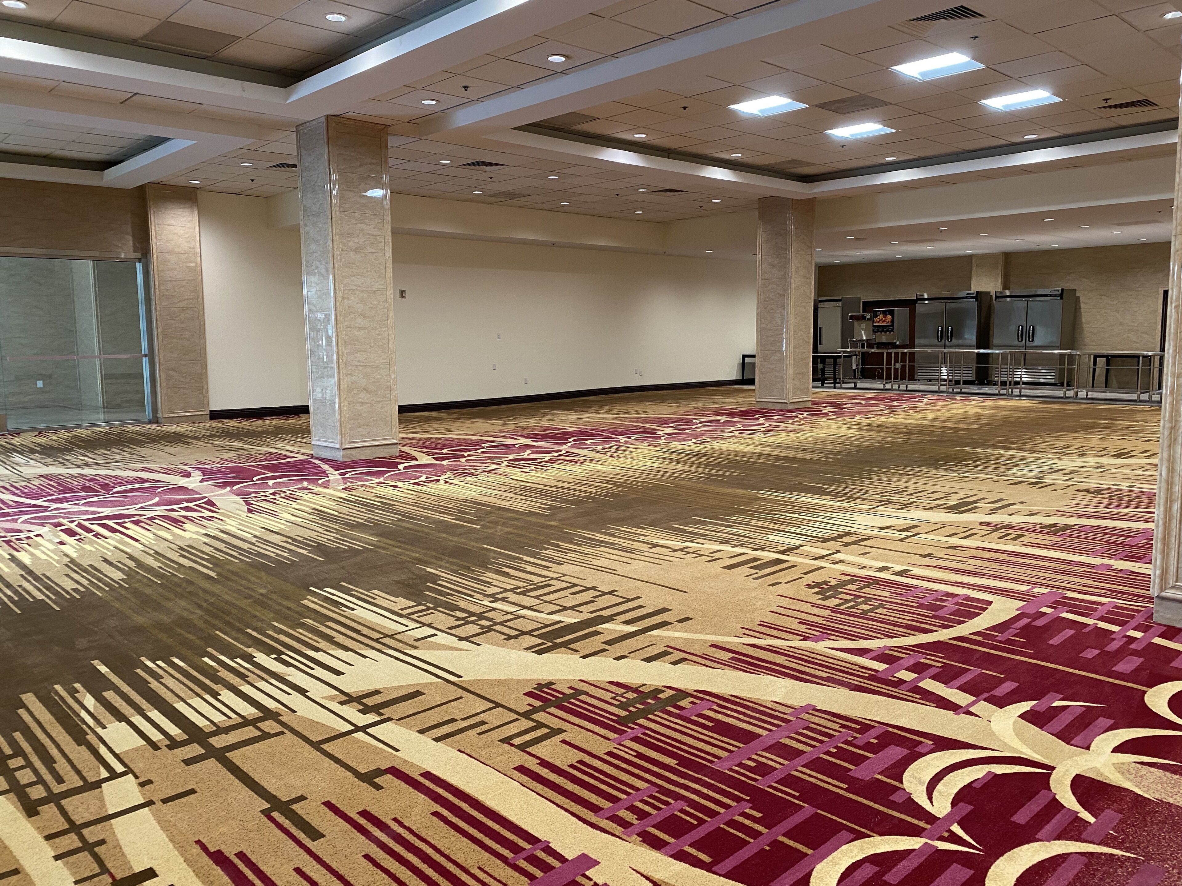 Um amplo hall de hotel com carpete estampado, pilares grandes e divisória de vidro.