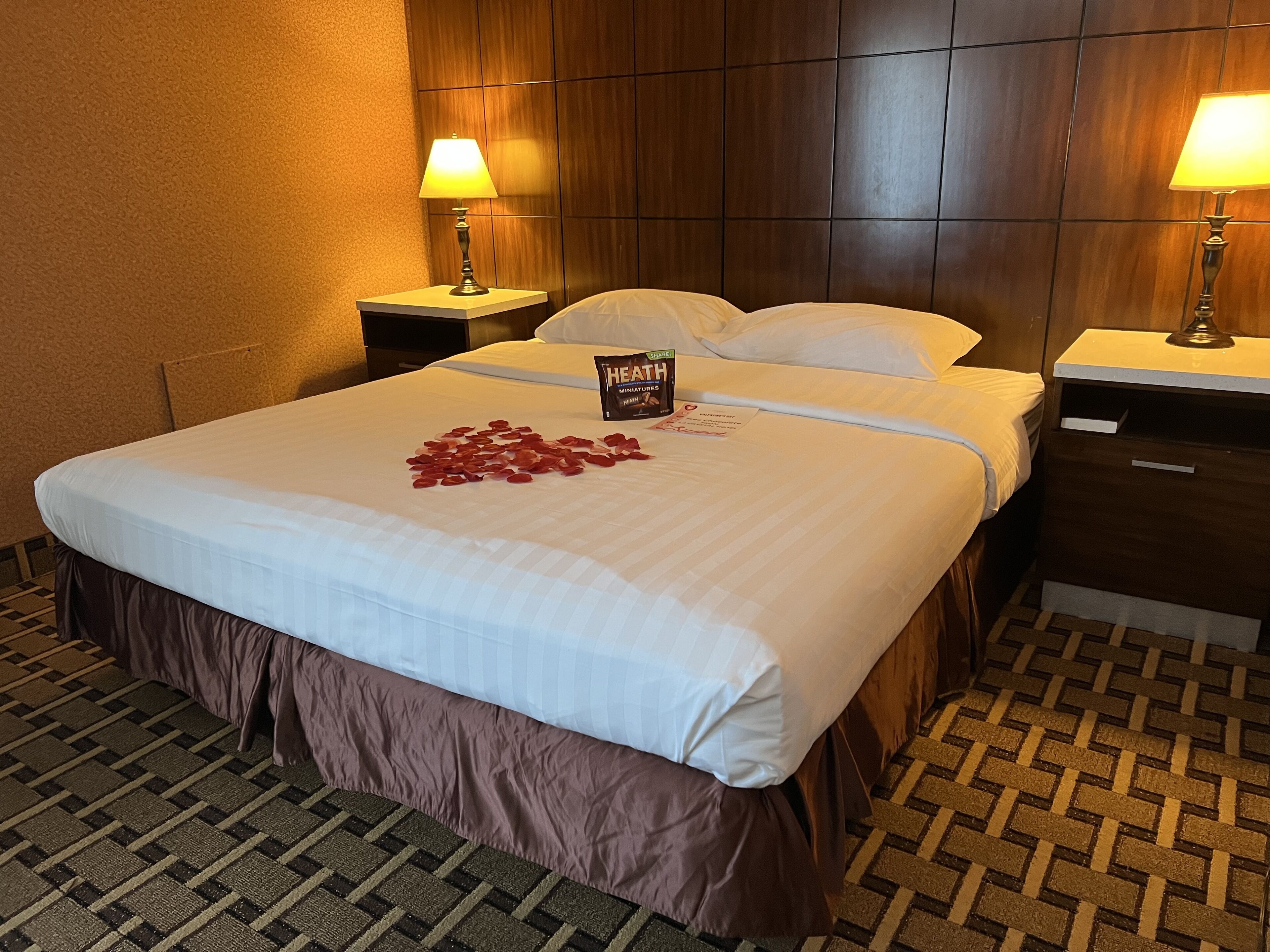 A hotel room with a neatly made bed, two bedside lamps, a wooden headboard, and a patterned carpet.