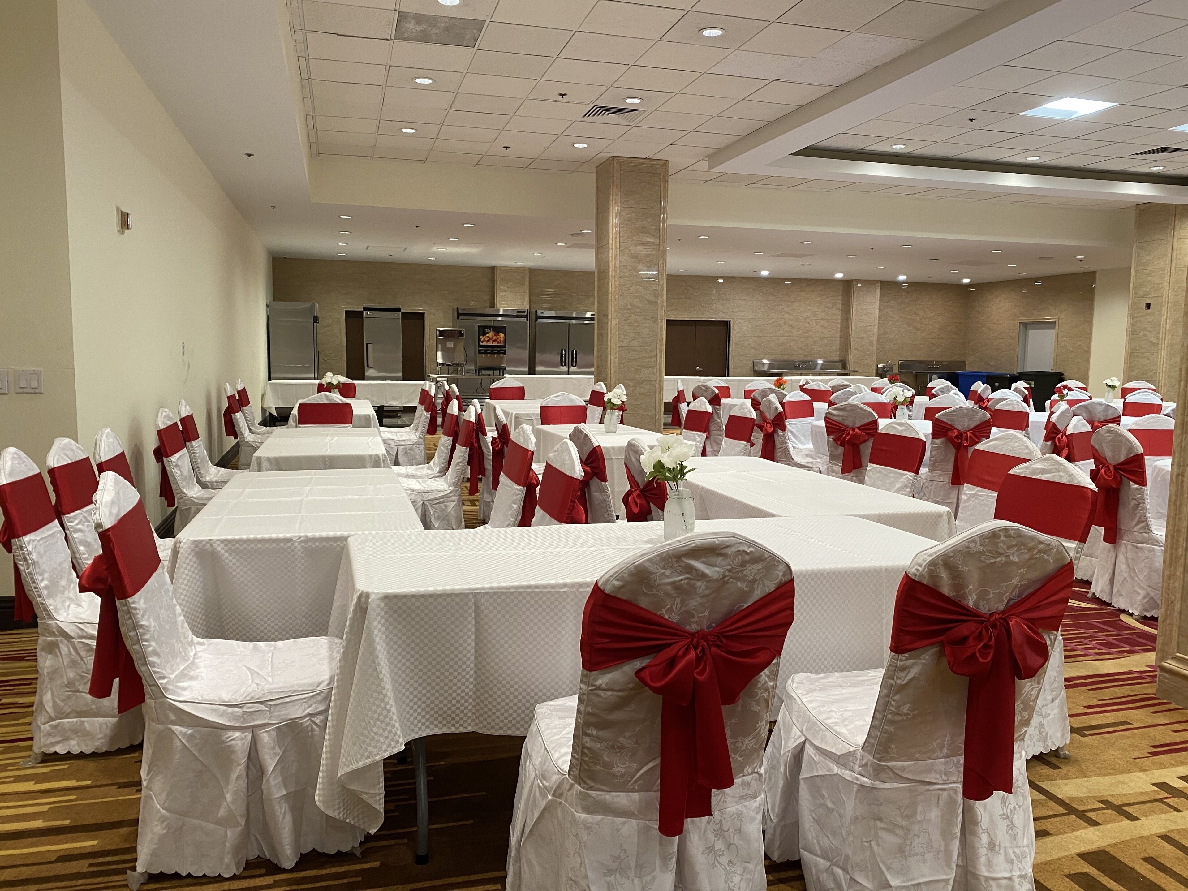 A banquet hall with rows of tables covered in white tablecloths, chairs with white and red sashes, and a central aisle.