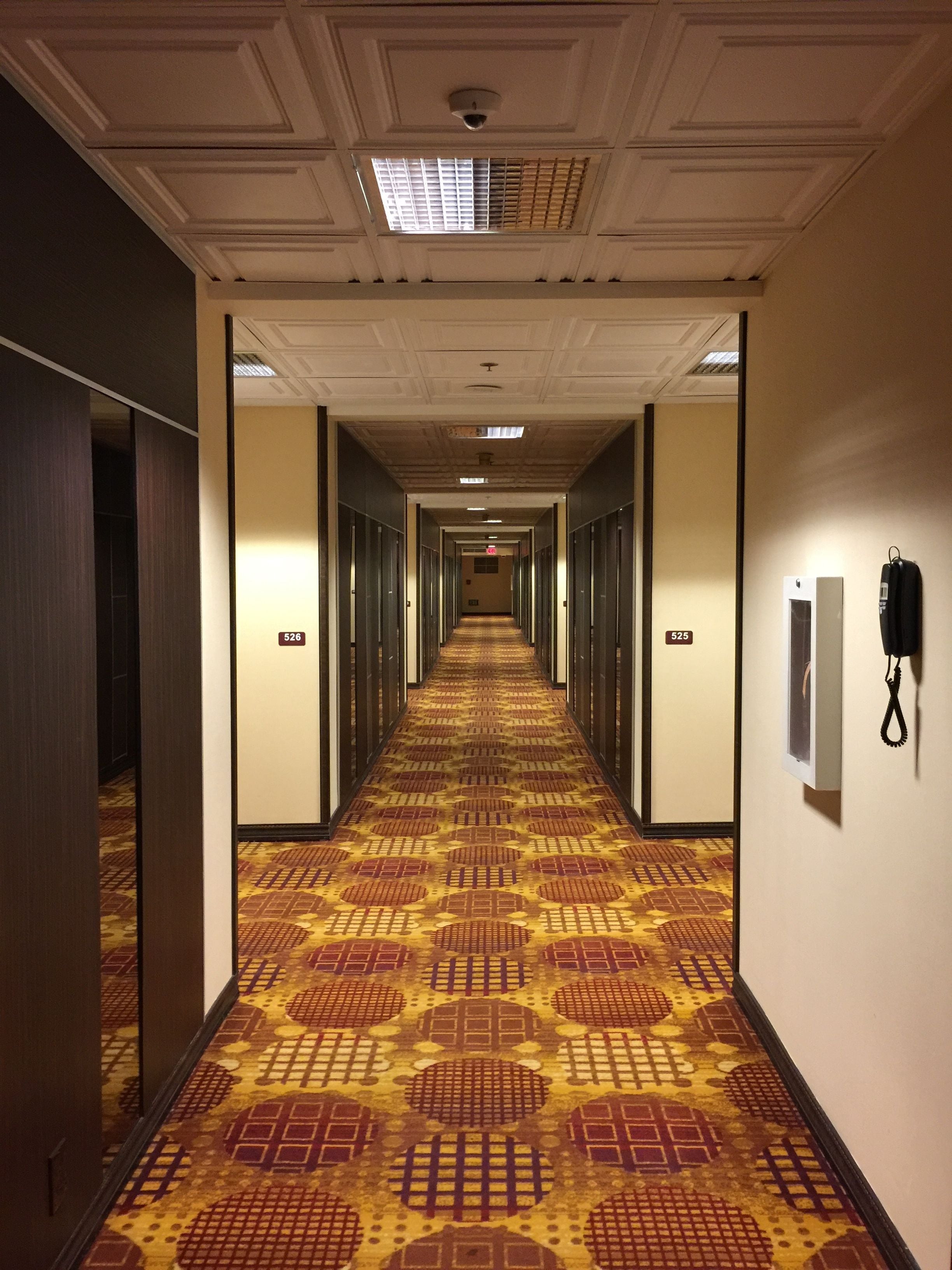 Un pasillo de hotel con alfombra estampada, puertas numeradas y una cámara de seguridad en el techo.