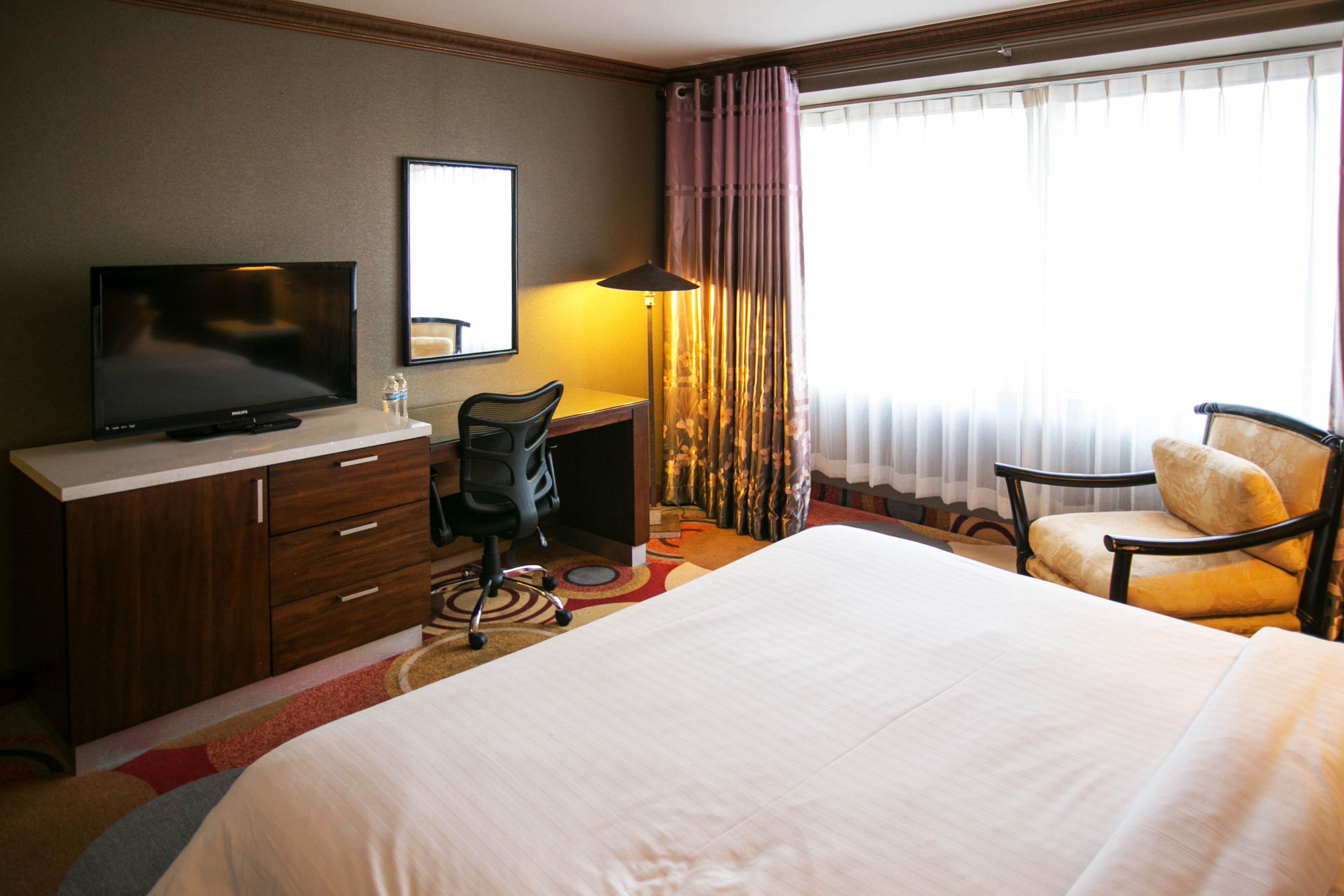 A hotel room with a bed, a desk with a television, a chair, and a window with curtains.