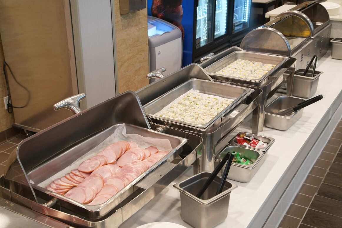 A buffet line with various food items, including sliced meats and sushi, and a refrigerator in the background.