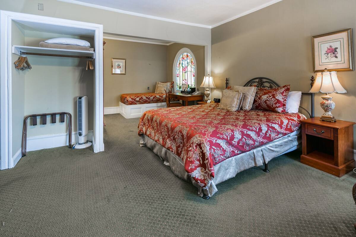 Quarto com uma cama grande, mesa de cabeceira com abajur, cadeira, escrivaninha, guarda-roupa e janela com vitrais.