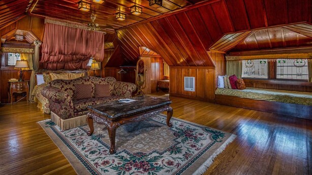 Un dormitorio ático acogedor con techo de madera, un sofá mullido, una mesa de centro de vidrio y un ventanal con cortinas.